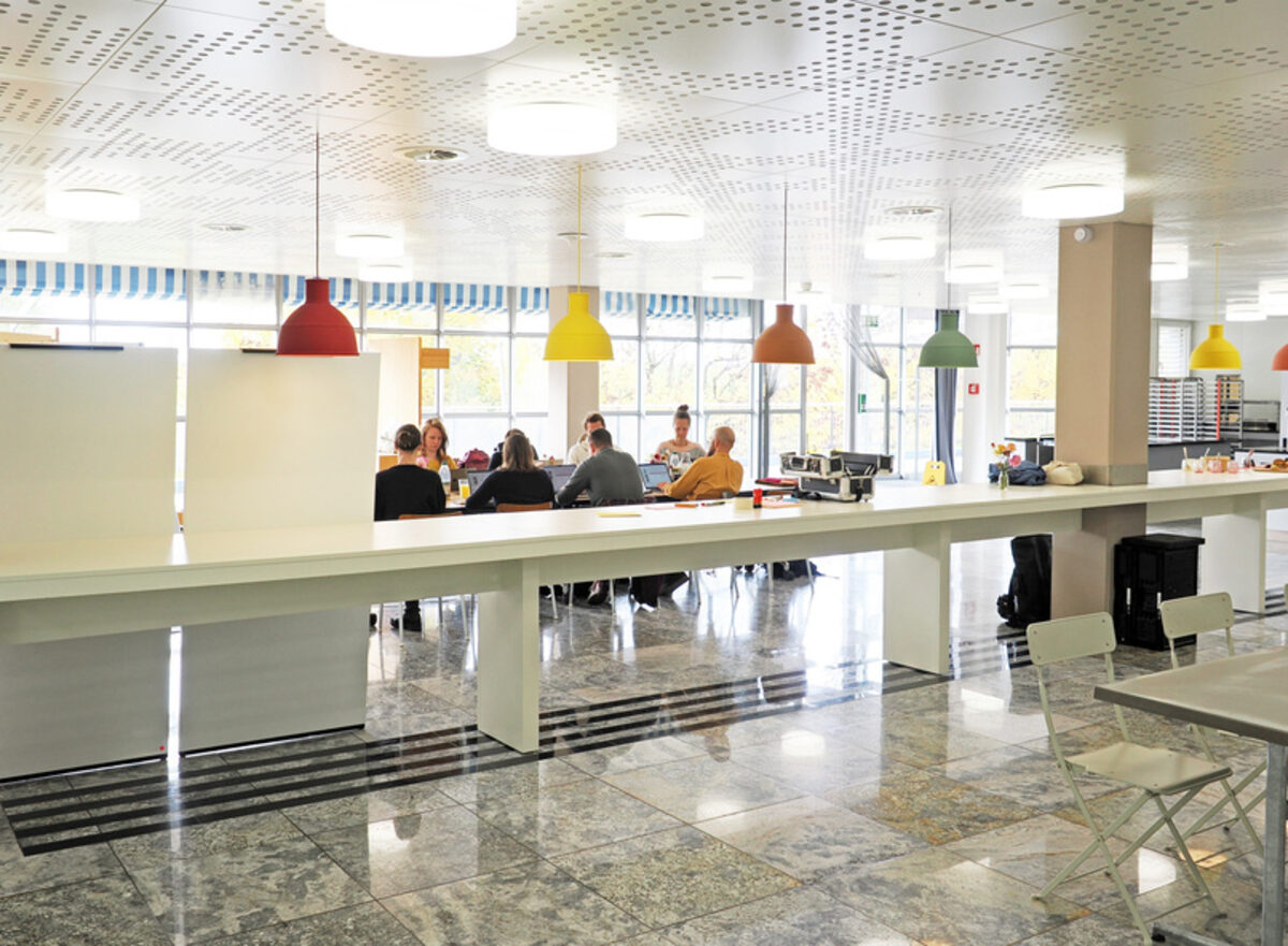 Heller Gemeinschaftsraum im RegioFood Hub mit langer Theke, farbigen Hängelampen und Menschen an Tischen.