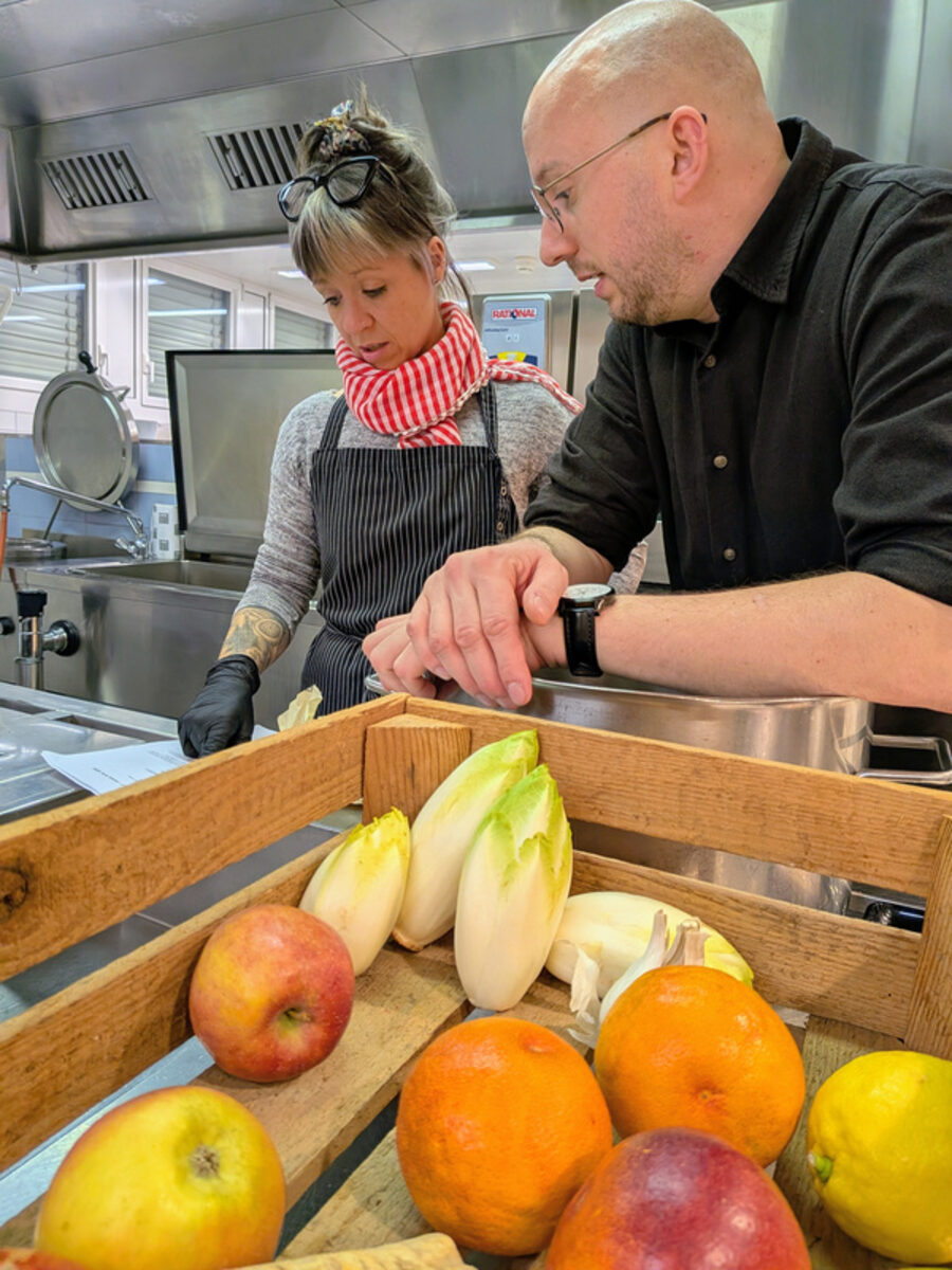 Dario Zbinden und eine Teilnehmerin schneiden in der Produktionsküche Gemüse, davor eine Harasse mit Früchten.