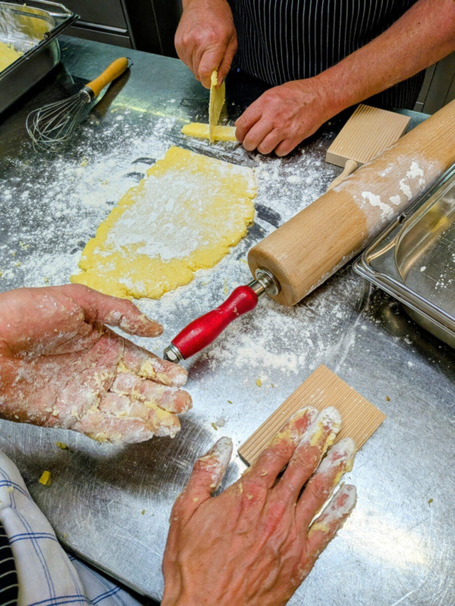 Vier bemehlte Hände formen auf einem Stahltisch Teig, daneben ein Wallholz mit rotem Griff.